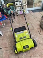 Front angled view showing bright green SunJoe lawn dethatcher with black handle and wheels on a stone patio.
