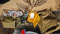 Photo showing fishing reels, tackle boxes with lures, small tool, and two beige canvas fishing bags on a carpet