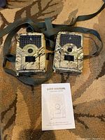 Two camouflaged game trail cameras lying flat with attached straps and a user manual placed below them, showing front view of the cameras.