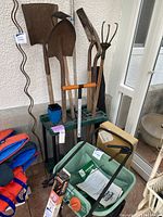 Photo shows various gardening tools standing against a textured white wall including shovels, spade, rake, lawn puncher, tomato stakes, and two green garden tool caddies (one in box). Fertilizer spreader and power rake are positioned in front.