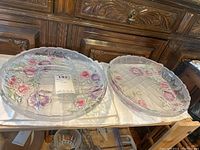 Two clear glass serving plates side by side on white surface, showing floral designs and scalloped edges.