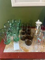 Set of green Coca-Cola glasses paired with smoky brown glasses and two labeled glass bottles, placed on a marble countertop.