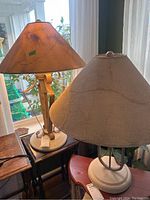 Two lamps placed indoors near window and other furniture. Both have concrete bases and beige fabric shades. One lamp has a slightly worn or stained lampshade.