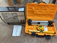 Full view of Johnson laser level kit in open orange case showing tripod, laser level, leveling bar, and instruction manual. Next to it is a Torcan portable heater.