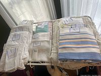 Multiple folded white linen luncheon napkins with embroidery and lace trims, folded striped napkins and tablecloths on top of crocheted linens.