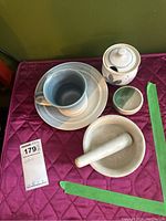 Overview photo of pottery set displayed on purple cloth with green background, showing cup and plate, mustard jar, small bowl, and white marble mortar with pestle.