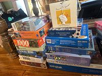 Stack of assorted puzzles including 1000, 750, 500 piece counts with different themes and brands visible on the boxes.