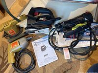 Black & Decker electric drill, Skil jigsaw, RotoZip Rebel spiral saw, sander, and Skil jigsaw manual on textured carpet
