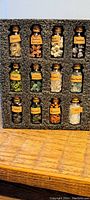 Photo showing 12 small glass bottles filled with various labeled gemstones arranged in black foam insert on wooden surface.