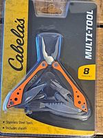 Front view of Cabela's Multi-Tool in original packaging showing orange and black handle, stainless steel tools, and label indicating 8 tools in 1.