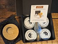 Top view of cocktail smoker kit inside box showing four tins labeled cherry, pecan, oak, and apple wood chips, wooden container, cleaning brush, chimney with steel mesh, and user manual.