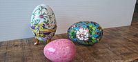 Three decorative eggs arranged on wooden surface with neutral background: white porcelain trinket egg with floral, blue cloisonné style egg, and pink stone egg.