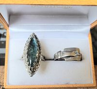Photo showing two fashion rings in a presentation box: one silver tone size 9 ring with a buckle detail, one adjustable ring with a blue elongated stone and ornamental setting.