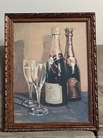 Framed print showing two champagne bottles and two champagne glasses on a table under glass with a wood frame. Minor wear on frame visible.