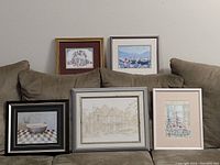 Five framed prints sitting on a couch, showing variety of art styles and frame materials, some matted, glass covered.