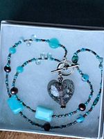 Colourful beaded necklace with turquoise, blue, and brown beads, glass heart pendant with purple and white details, silver-tone toggle clasp, displayed on light-colored box.