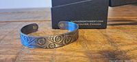 Silver-toned cuff bracelet with multiple etched hummingbird motifs arranged in repeating pattern. Bracelet shown on wooden surface with black Native Northwest box behind.