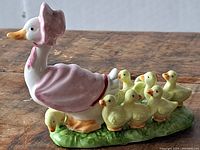 Side view showing mother goose in pink bonnet and cape leading seven yellow baby geese on green grass base.