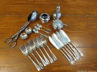 Photo showing the entire lot of silver plated utensils on a wooden surface including forks, knives, tongs, small spoons, salt cellar with spoon, sugar bowl, and pepper shaker.