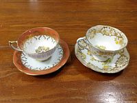 Two tea cups and two saucers side by side on a wooden surface, showing the Royal Albert set with coral and gold design and the Aynsley set with yellow floral and gold accents.