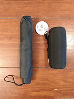 Two black compact umbrellas side by side on wooden floor, one with fabric cover and one in black zippered case.