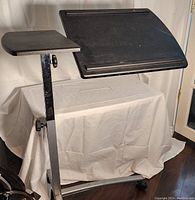 Photo of the adjustable black bed table showing two platforms, one tilt adjustable large platform and one smaller flat side platform, with height adjustment and caster wheels.