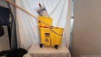 Photo shows yellow commercial mop bucket with inner bucket, mounted on four black caster wheels, wooden handle mop inserted in bucket, replacement mop head in packaging leaning on bucket.