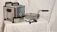 T-fal stainless steel deep fryer with basket, glass lid, oil filter, and lower oil storage bin shown on white tablecloth background.