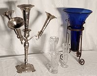 Photo showing a silver epergne with three fluted trumpet arms, large cobalt blue vase on scroll metal stand, and three clear glass bud vases of varied shapes and patterns.