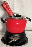 Photo showing red ceramic fondue pot on black stand with fuel pot and red-handled fondue forks inside the pot.