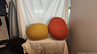 Featured two color-knit poufs, one yellow and one orange, placed on a white surface in front of a white curtain backdrop.