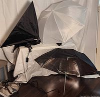Two reflective umbrellas (one black and silver interior, one white translucent) and one opened square reflective umbrella with light socket shown upright on floor and table.