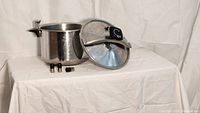 Photo shows the entire pressure cooker set including the large stainless steel pot, the lid with built-in pressure gauge, and three different metal pressure weights beside it.