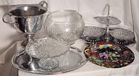 Photo showing a silver-plated trophy bowl with handles, oval serving tray, large engraved glass bowl, glass pedestal cake stand with textured pattern, two-tier segmented silver-plated serving tray, two divided etched glass serving dishes, and a colorful decorative plate.