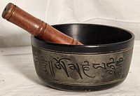 Photo of black metal singing bowl with engraved script on the outer side, along with a cylindrical wooden striker resting inside the bowl.