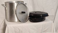 Photo showing an aluminum stock pot with lid on left and a black enamel roaster with lid stacked on right, placed on a white cloth background.