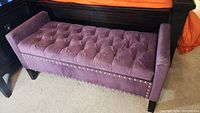 View of the tufted purple velour storage bench closed, showing nailhead trim and black wooden legs.
