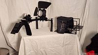 Teleprompter and shoulder rig shown on a white draped table from a front angle. Teleprompter has a black hood and glass. Shoulder rig metal frame has foam hand grips.