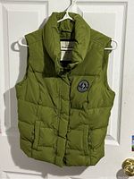Green sleeveless puffer vest with front snap buttons, side pockets, and logo patch on chest, hanging on a white clothes hanger in front of a white door.