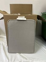 Front view of one grey outdoor chair cushion propped against a box showing rectangular shape with piped edges.