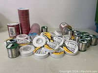 Wide view showing multiple solder wire spools and electrical tape rolls mostly in red, white and metallic green spools on a surface.