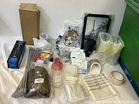 Photo showing large lot of household and kitchen items on white cloth including various cleaning tools, zip ties, disposable gloves, and framed photo.