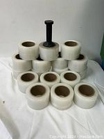 Seventeen rolls of clear stretch wrap stacked in a pyramid with a single hand-held handle placed on top of the center roll.