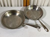 Two All-Clad stainless steel skillets (8 and 7 inch) placed side by side on a white surface.