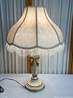 Classic Mid Century Modern table lamp with ivory and gold painted metal base and ivory silk lampshade with fringe trim, on a white surface against a curtain.