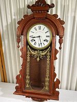 Full front view showing wooden carved case, glass door with gold decoration, dial with Roman numerals and brand name.