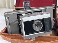 Polaroid Land Camera Model J66 open with folding bellows extended, showing front camera lens, light adjustment slider, and built-in viewfinder, inside brown leather case.