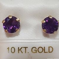 Close-up of pair of 10KT yellow gold stud earrings with round amethyst stones in prong settings on a white textured background.