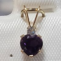 Close-up front view of heart-shaped purple amethyst set in yellow gold with small clear accent stone and gold bail.