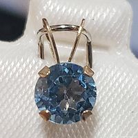 Close-up image showing round blue topaz gemstone in 10K yellow gold setting with four prongs and bail attachment.